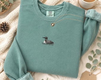 May include: A sage green crewneck sweatshirt featuring a small embroidered loon on the center chest. The garment has a ribbed collar, cuffs, and hem, with a Comfort Colors brand label visible at the inner neckline. The fabric has a soft, matte finish.
