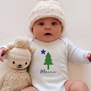 May include: A white baby onesie with a green pine tree and a blue star, and the word "Maine" printed on the front. A baby is wearing the onesie and holding a beige plush toy bear.