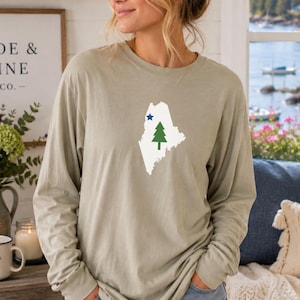 Könnte beinhalten: Ein langärmeliges Rundhals-Shirt in einem gedämpften Salbeigrün. Auf der Brust befindet sich eine weiße Grafik, die den Umriss des Bundesstaates Maine darstellt, mit einem kleinen blauen Stern oben links und einer grünen Tanne in der Mitte.