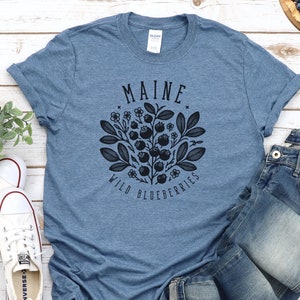 May include: A blue t-shirt with a graphic design of a blueberry bush and the text "Maine Wild Blueberries". The shirt is laid out on a white wooden surface with a pair of white sneakers, blue jeans, and a plate of blueberries.
