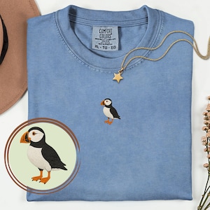 Könnte beinhalten: Hellblaues Comfort Colors T-Shirt mit einem kleinen, gestickten Papageientaucher. Der Papageientaucher hat einen schwarzen Rücken, einen weißen Bauch, orangefarbene Füße und einen großen orange-gelben Schnabel. Eine goldene Sternanhänger-Halskette und ein brauner Hut sind ebenfalls im Bild.