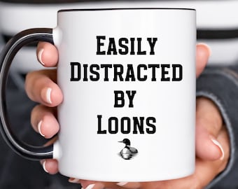 May include: White ceramic mug with a black rim and handle. The mug has black text that reads "Easily Distracted by Loons" with a black loon illustration below the text.