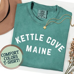 May include: Teal Comfort Colors t-shirt with "Kettle Cove Maine" printed in white. A gold star pendant necklace and a brown hat are also visible. The shirt has a round neck and short sleeves. A round sticker says "Comfort Colors T-Shirt!"