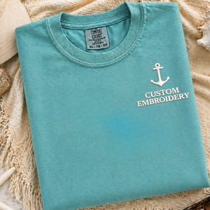 May include: A turquoise Comfort Colors t-shirt with a white anchor and the words "CUSTOM EMBROIDERY". The shirt is folded and lies on a beige surface. A hat, seashells, and sunglasses are in the background.
