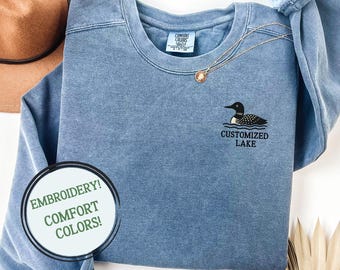 Puede incluir: Una sudadera azul de cuello redondo con una etiqueta Comfort Colors en el interior del cuello. El pecho presenta un gráfico bordado de un colimbo en blanco y negro sobre el texto "CUSTOMIZED LAKE". Una cadena dorada descansa sobre la tela.