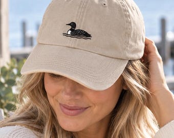 Può includere: Un cappellino da baseball beige con un ricamo nero e bianco raffigurante una strolaga sulla parte anteriore. Il cappellino presenta una visiera curva, una corona strutturata con occhielli di ventilazione e una chiusura in tessuto sul retro.
