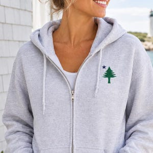 May include: A light gray zip-up hoodie featuring a small embroidered green pine tree graphic with a dark blue star positioned above the tree on the left chest area. The hoodie includes a front metal zipper, a hood with drawstrings, and two pockets.