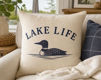 Può includere: Un cuscino quadrato con una fodera in tessuto testurizzato color crema. La parte anteriore presenta una grafica blu scuro di un colimbo che nuota sull'acqua, posizionata sotto la scritta "LAKE LIFE" stampata in un carattere serif blu.