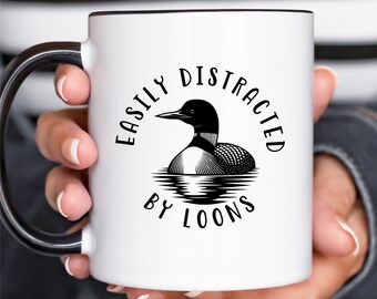 May include: A white ceramic mug with a black handle and rim. The front features a black graphic illustration of a loon swimming on water, surrounded by the text "EASILY DISTRACTED BY LOONS" printed in a bold, black, sans-serif font.