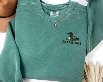 Puede incluir: Una sudadera de color verde azulado con el texto "ON LAKE TIME" y un gráfico de un somorgujo. Un collar de oro con un colgante está drapeado sobre la sudadera. Un sombrero marrón es visible en la esquina superior izquierda.