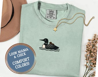 May include: A light green Comfort Colors t-shirt featuring an embroidered design of a loon and its chick on water. The shirt has a crew neck and short sleeves. A gold star pendant necklace and a brown hat are also in the image. The text "LOON MAMA & CHICK COMFORT COLORS!" is in a circle.