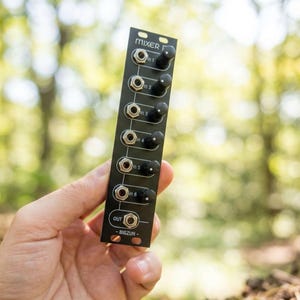 Puede incluir: Un módulo mezclador de audio negro con la palabra "MIXER" en la parte superior y "BIGZUN" en la parte inferior. Tiene seis conectores de entrada, seis perillas y un conector de salida. El módulo se sostiene en una mano contra un fondo verde borroso.