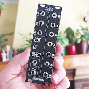 Peut inclure: Un module de synthétiseur modulaire Eurorack noir étiqueté Clock Divider et Out Of Even. La façade présente plusieurs prises d'entrée et de sortie en métal, des étiquettes blanches pour les divisions et un logo Bigzun en bas. Il a des trous de montage.