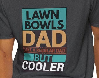 May include: Dark gray t-shirt with the text "LAWN BOWLS DAD LIKE A REGULAR DAD BUT COOLER" in teal, tan, and white. The shirt is a crew neck and is being worn.