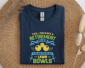 May include: A navy blue t-shirt with a graphic print in yellow, light blue, and lime green. The text reads: YES, I DO HAVE A RETIREMENT PLAN I PLAN TO PLAY LAWN BOWLS, accompanied by an illustration of three yellow lawn bowls.
