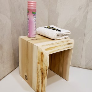 May include: A wooden stool with a natural wood finish and a curved design. The stool is made from a single piece of wood and has a unique grain pattern. A pink can of shaving cream and a white towel are on top of the stool.