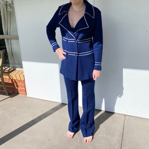 May include: A navy blue pantsuit with white piping. The jacket has a double-breasted closure and a collar with a pointed lapel. The trousers have a straight leg.