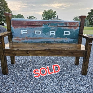 May include: A rustic wooden bench with a weathered blue and green Ford tailgate as the backrest. The bench has a dark brown stain and is sitting on a gravel surface. The words "SOLD" are written in red on the front of the bench.