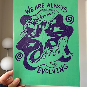 Puede incluir: Impresión verde con un diseño morado de dos ranas y el texto "We are always evolving" con un diseño de remolino.