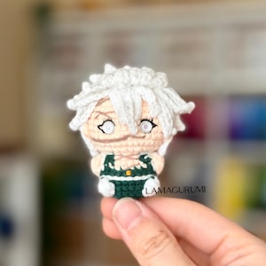 Op de afbeelding: Handgemaakte gehaakte pop met wit haar en een groene outfit. De pop heeft een gedetailleerd gezicht met geborduurde kenmerken. De pop is een klein, handgemaakt item, waarschijnlijk een amigurumi. De pop wordt in een hand gehouden.