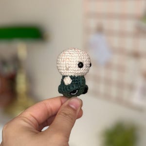 May include: A small crocheted figure with a white head and green clothing. The figure has black eyes and a simple, rounded shape.