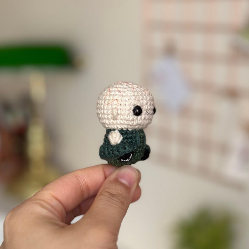 May include: A small crocheted figure with a white head and green clothing. The figure has black eyes and a simple, rounded shape.