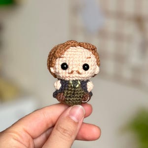 Puede incluir: Una muñeca amigurumi de crochet de un hombre con cabello castaño, bigote y un atuendo negro y verde.