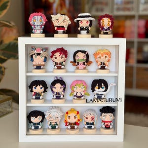 May include: A white wooden display shelf containing sixteen small crocheted character figurines arranged on four levels. Each figure stands on a circular wooden base. The text LAMAGURUMI is printed in black on the bottom right shelf area.