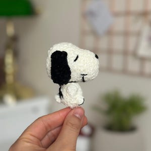 May include: A handmade, white and black Snoopy amigurumi. The plush toy features Snoopy's signature black ears, black spot, and black collar. The amigurumi is made of a soft, textured yarn.