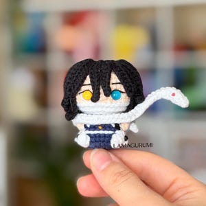 Könnte beinhalten: Eine kleine gehäkelte Amigurumi-Figur mit schwarzem Haar, einem gelben und einem blauen Auge. Die Figur trägt eine weiße Gesichtsbedeckung und eine weiße Schlange um den Hals. Der Körper ist dunkelblau und weiß. Der Text LAMAGURUMI ist sichtbar.