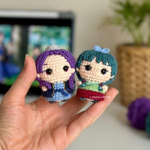 Puede incluir: Dos pequeñas muñecas de ganchillo hechas a mano. Una muñeca tiene el pelo morado y un atuendo azul, la otra tiene el pelo verde azulado y un atuendo verde y rojo. Las muñecas tienen ojos negros y se sostienen en una mano. El texto "LAMAGURUMI" es visible en la segunda muñeca.