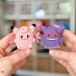 Könnte beinhalten: Zwei kleine gehäkelte Figuren, bestehend aus einem hellrosa Charakter mit schwarz gespitzten Ohren und einem violetten Charakter mit roten Augen und einem breiten weißen Grinsen. Der violette Charakter trägt den weißen Schriftzug LAMAGURUMI.