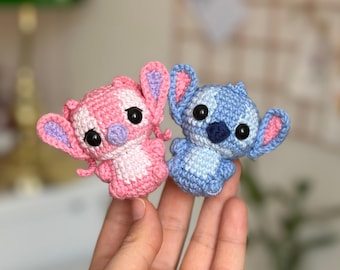 May include: Two crocheted figures of the character Stitch, one pink and one blue. The pink figure has a white and pink pattern, while the blue figure is solid blue. Both have large black eyes and large ears.
