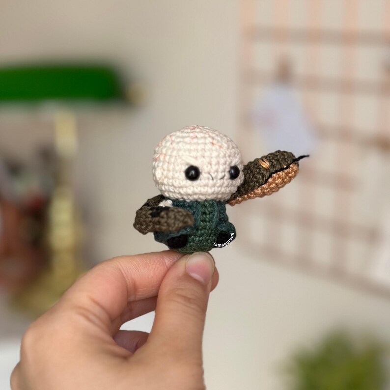 May include: A crocheted amigurumi figure of a bald man with a snake wrapped around his arm. The man is wearing a green shirt and the snake is brown and green.