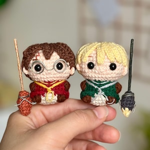 Puede incluir: Dos figuras de amigurumi de crochet de Harry Potter y Ron Weasley, cada uno sosteniendo una escoba. Harry lleva una túnica roja y dorada y gafas redondas. Ron lleva una túnica verde.
