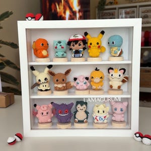 Puede incluir: Un estante blanco que contiene quince pequeñas figuras de ganchillo de varias criaturas, cada una montada sobre una base de madera circular. La colección incluye personajes como Pikachu, Charmander y Gengar. El texto LAMAGURUMI es visible.