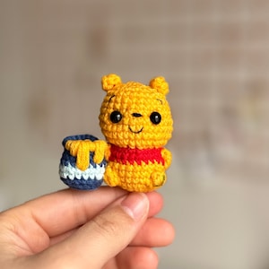 May include: A small crocheted Winnie the Pooh plush toy holding a blue and white honey pot. The toy is yellow with a red shirt and black eyes.