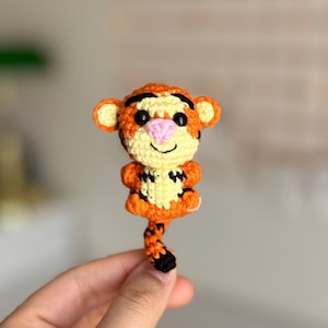 May include: A small, crocheted tiger figurine in orange, black, and cream colors. The tiger has a pink nose, black eyes, and a smiling face. The tiger's tail is striped and black at the tip. The figurine is a handmade item.