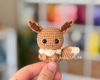 May include: A small, brown, crocheted amigurumi figure resembling a fox-like creature with large black eyes, pointed ears with dark brown centres, and a cream-coloured fluffy collar. The text LAMAGURUMI is visible on the lower right side.