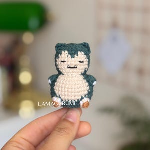 Könnte beinhalten: Ein kleines, dunkelblaugrünes und cremefarbenes Häkel-Plüschtier in Form einer Snorlax-Figur. Die Figur hat geschlossene Augen, einen kleinen Mund mit zwei sichtbaren Zähnen und abgerundete Gliedmaßen mit weißen Akzenten. Der Text LAMAGURUMI ist darunter aufgedruckt.