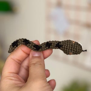 May include: A small crocheted snake with black and green stripes and brown eyes. The snake is being held by a hand.