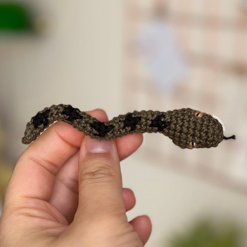 May include: A small crocheted snake with black and green stripes and brown eyes. The snake is being held by a hand.