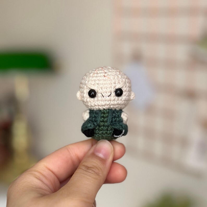 May include: A crocheted amigurumi toy of a bald man with a green robe and a mischievous smile. The toy is small and is being held in a hand.
