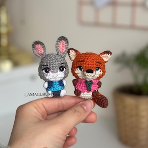 May include: Two crocheted amigurumi figures: a gray bunny with pink-lined ears and a fox with orange fur. The bunny wears a blue vest, and the fox wears a pink shirt and green tie. The figures are held in a hand.