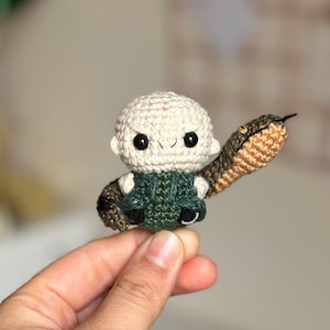 Puede incluir: Un juguete de amigurumi tejido a crochet de un personaje con una serpiente. El personaje es blanco con ojos negros y una camisa verde. La serpiente es marrón y naranja.