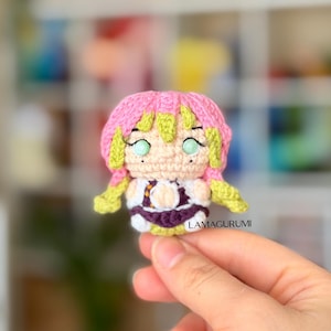 May include: A small crochet doll featuring pink and lime green hair, light skin, and large light green eyes. The figure wears a dark purple and white outfit with orange accents. The text LAMAGURUMI is printed in black on the lower right side.