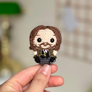 May include: A crocheted amigurumi doll of a man with brown hair, a brown beard, and a mustache. He is wearing a green jacket, a black vest, a white shirt, and gray pants.