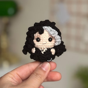 May include: A handmade crochet doll with black hair, a gray streak, and a black outfit. The doll has a smiling face with black eyes and is held in a hand. The doll is a small, handcrafted item.