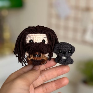 May include: Two crocheted amigurumi figures: a brown-haired man with a beard and a black dog. The man is wearing a brown and tan outfit and the dog has white accents on its face.