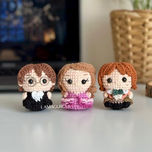 Puede incluir: Tres muñecas de ganchillo hechas a mano con cabello castaño, castaño claro y rojo. Las muñecas visten trajes negros, rosas y marrones. La muñeca de la izquierda tiene gafas. Las muñecas están sobre una superficie blanca. El texto "LAMAGURUMI" es visible.